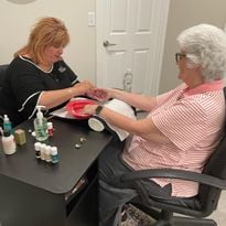 Two women face each other over a manicure table, as one woman gives the other woman a manicure.